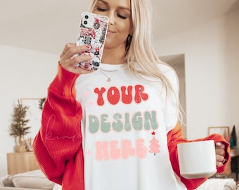 May include: White t-shirt with a red, green, and pink design that says "YOUR DESIGN HERE" with a small red star and a green Christmas tree. The shirt is being worn by a person with blonde hair and a red cardigan.