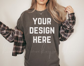 Puede incluir: Una camiseta gris con el texto "YOUR DESIGN HERE" impreso en blanco en la parte delantera. La camiseta la lleva puesta una persona con el pelo largo rubio y una camisa de cuadros.