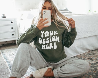 May include: A woman wearing a green sweatshirt with the text "YOUR DESIGN HERE" in white. She is sitting on a rug with her legs crossed and holding a phone to her face.