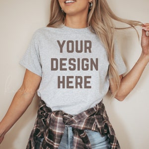 Puede incluir: Una camiseta gris con el texto "YOUR DESIGN HERE" impreso en marr&oacute;n en la parte delantera. La camiseta la lleva puesta una persona con el pelo largo y rubio y est&aacute; atada a la cintura con una camisa de cuadros.