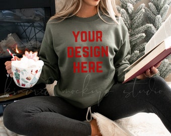 May include: A woman wearing a green sweatshirt with the text "YOUR DESIGN HERE" in red. She is sitting on a white fuzzy rug and holding a book and a mug.