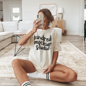 May include: A woman wearing a white t-shirt with the text "kindred mockup studio" in black lettering. She is sitting on a light brown rug with her legs crossed and wearing white socks with black stripes.