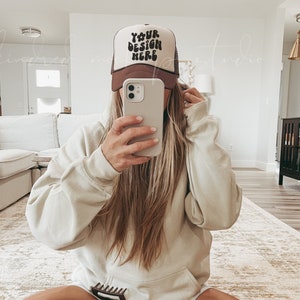 May include: A person wearing a brown and white trucker hat with the text "YOUR DESIGN HERE" in black. The person is wearing a light beige hoodie and white socks with black stripes. The person is sitting on a rug with their legs crossed.