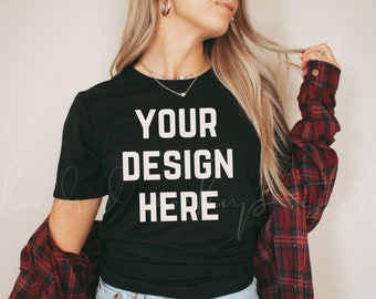 以下が含まれることがあります： 白い文字で「YOUR DESIGN HERE」と書かれた黒いTシャツ。長いブロンドの髪と赤いチェックのシャツを着た人が着ています。