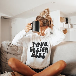 May include: A white long-sleeve sweatshirt with the text "YOUR DESIGN HERE" in black, retro-style lettering. The sweatshirt is being worn by a person sitting on a couch.