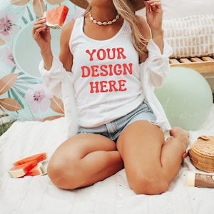 Op de afbeelding: Een wit tanktop met de tekst "YOUR DESIGN HERE" in rood. Het tanktop wordt gedragen door een persoon die op een wit deken zit.
