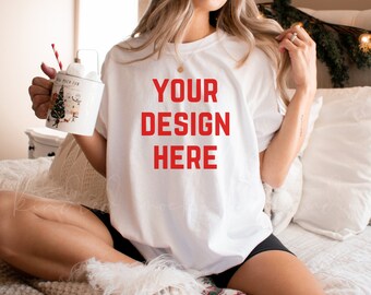 May include: A white t-shirt with the text "YOUR DESIGN HERE" in red letters. The shirt is being worn by a person sitting on a bed with a white blanket.