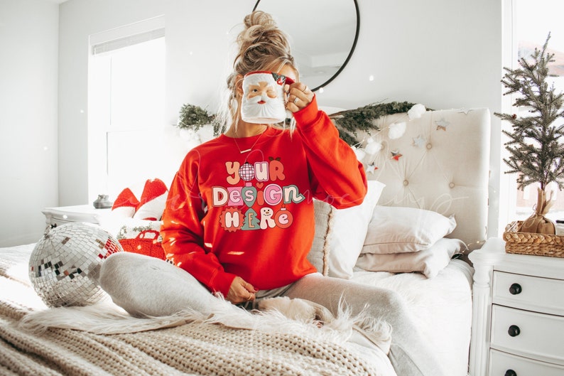 May include: A woman wearing a red sweatshirt with the text "YOUR DESIGN HERE" in colorful letters. She is holding a white mug with a Santa Claus face on it. The woman is sitting on a bed with white bedding and a white headboard. There is a small Christmas tree in the background.