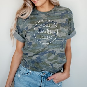 May include: A woman wearing a green, blue, and brown camouflage t-shirt with a white circle that says "your design here". She is wearing blue jeans and has her hand in her pocket.