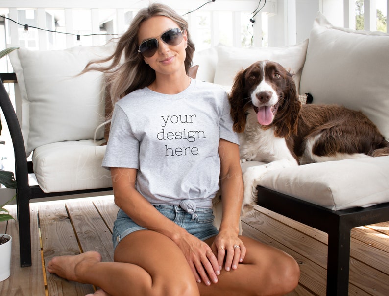 Peut inclure: Une femme portant un t-shirt gris avec le texte "your design here" assise sur une terrasse en bois avec un chien brun et blanc.