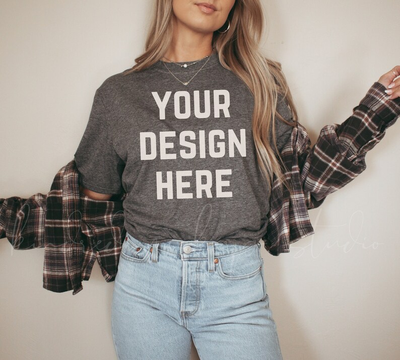 May include: A gray t-shirt with the text "YOUR DESIGN HERE" printed in white. The shirt is being worn by a person with long blonde hair and is paired with a plaid shirt and blue jeans.