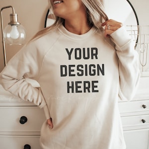 以下が含まれることがあります： クルーネックのロングスリーブのクリーム色のスウェットシャツ。スウェットシャツには、「YOUR DESIGN HERE」という文字が前面に印刷されています。