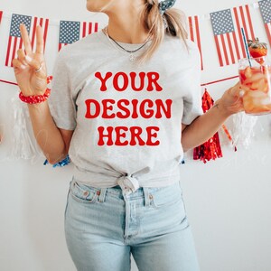 May include: A woman wearing a grey t-shirt with the text "YOUR DESIGN HERE" in red letters. She is holding a glass of red liquid with a straw and a strawberry garnish. The background is a white wall with a red, white and blue garland.