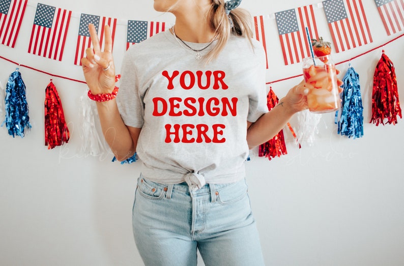 May include: A woman wearing a grey t-shirt with the text "YOUR DESIGN HERE" in red letters. She is holding a glass of red liquid with a straw and a strawberry garnish. The background is a white wall with a red, white and blue garland.