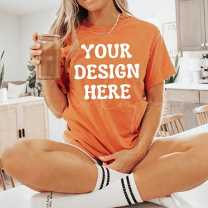 May include: A woman wearing a bright orange t-shirt with the text "YOUR DESIGN HERE" in white. She is sitting on a white surface with her legs crossed and holding a glass jar.