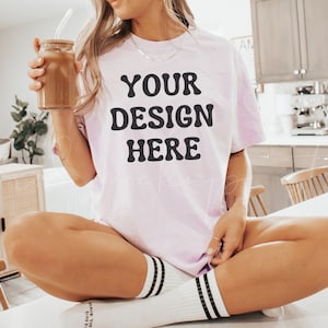May include: A woman wearing a light pink t-shirt with the text "YOUR DESIGN HERE" in black lettering. She is sitting on a white countertop with her legs crossed and holding a glass jar with a straw.