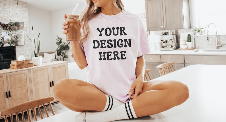 May include: A woman wearing a light pink t-shirt with the text "YOUR DESIGN HERE" in black lettering. She is sitting on a white countertop with her legs crossed and holding a glass jar with a straw.