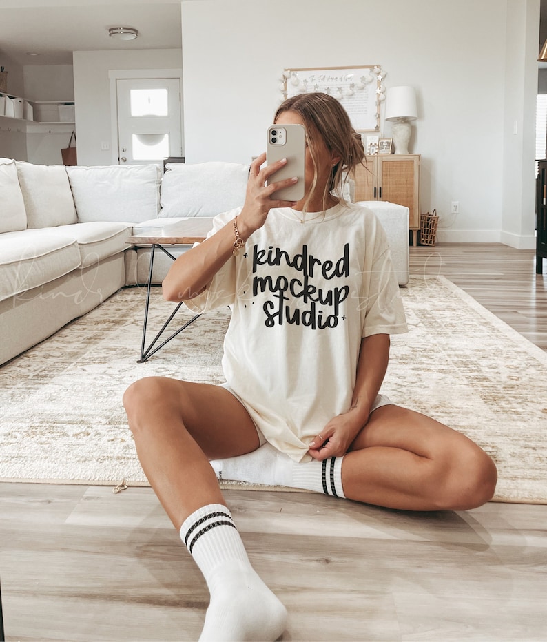 May include: A woman wearing a white t-shirt with the text "kindred mockup studio" in black lettering. She is sitting on a light brown rug with her legs crossed and wearing white socks with black stripes.