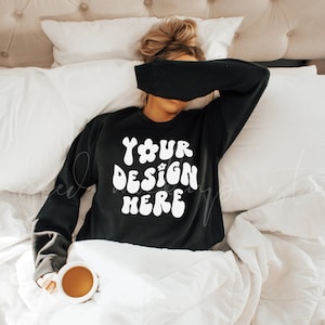 May include: A woman wearing a black sweatshirt with the text "YOUR DESIGN HERE" in white, lying in bed with a white blanket and a cup of coffee.