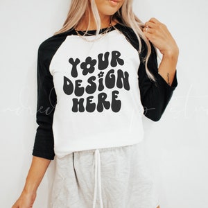 Op de afbeelding: Een wit en zwart baseballshirt met een zwarte afbeelding waarop in een retro-lettertype "YOUR DESIGN HERE" staat.