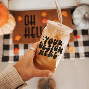 Op de afbeelding: Een glazen pot met een houten deksel en een rietje, gevuld met een bruine vloeistof. De pot heeft een zwart-wit ontwerp met de tekst "YOUR DESIGN HERE". De pot wordt vastgehouden door een hand voor een deurmat met een herfstthema.