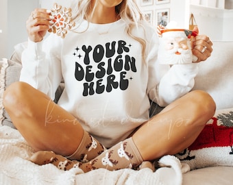 Puede incluir: Sudadera blanca con el texto "YOUR DESIGN HERE" en negro. La persona sostiene una galleta de copo de nieve y una taza de Papá Noel. Los calcetines tienen un estampado de renos. El tema es Navidad e invierno.