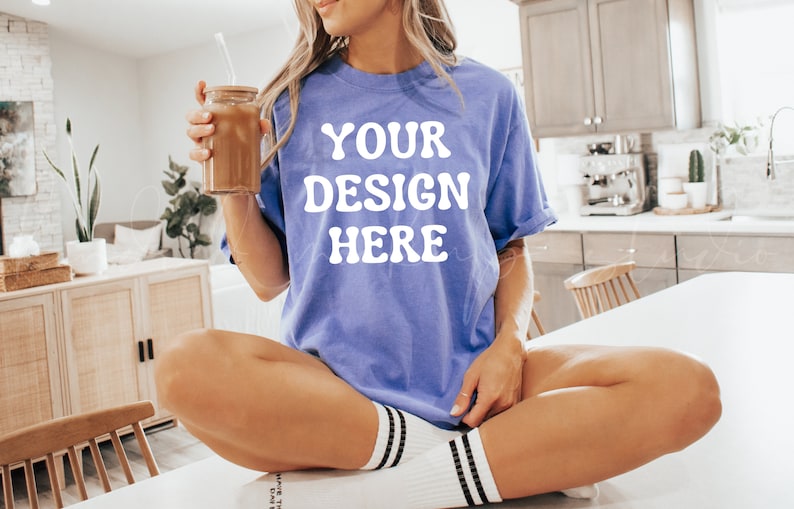 May include: A woman wearing a light purple t-shirt with the text "YOUR DESIGN HERE" in white. She is sitting on a white surface with her legs crossed and holding a glass with a straw.