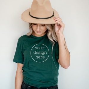 Puede incluir: Una mujer con una camiseta verde oscuro con el texto "your design here" en un círculo. Lleva un sombrero fedora marrón y pantalones vaqueros negros.