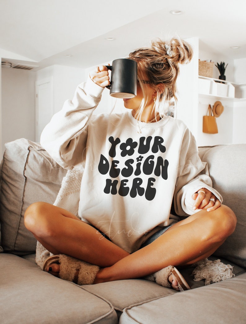 Peut inclure: Une femme portant un sweat-shirt blanc avec le texte "YOUR DESIGN HERE" en noir, assise sur un canap&eacute; beige, les jambes crois&eacute;es. Elle tient une tasse noire dans sa main droite.