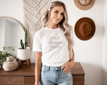 May include: A white t-shirt with the text "your design here" printed on the front. The shirt is being worn by a person with long blonde hair. The person is standing in front of a white wall with a macrame wall hanging and two brown hats.