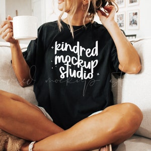 Op de afbeelding: Een vrouw draagt een zwart T-shirt met de tekst "kindred mockup studio" in witte letters. Ze zit op een bank en houdt een witte mok vast.