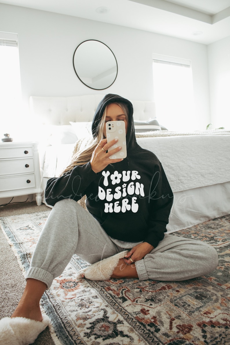 Puede incluir: Una sudadera con capucha negra con el texto "YOUR DESIGN HERE" en blanco. La modelo lleva la sudadera y pantalones de ch&aacute;ndal grises, sentada en una alfombra estampada.