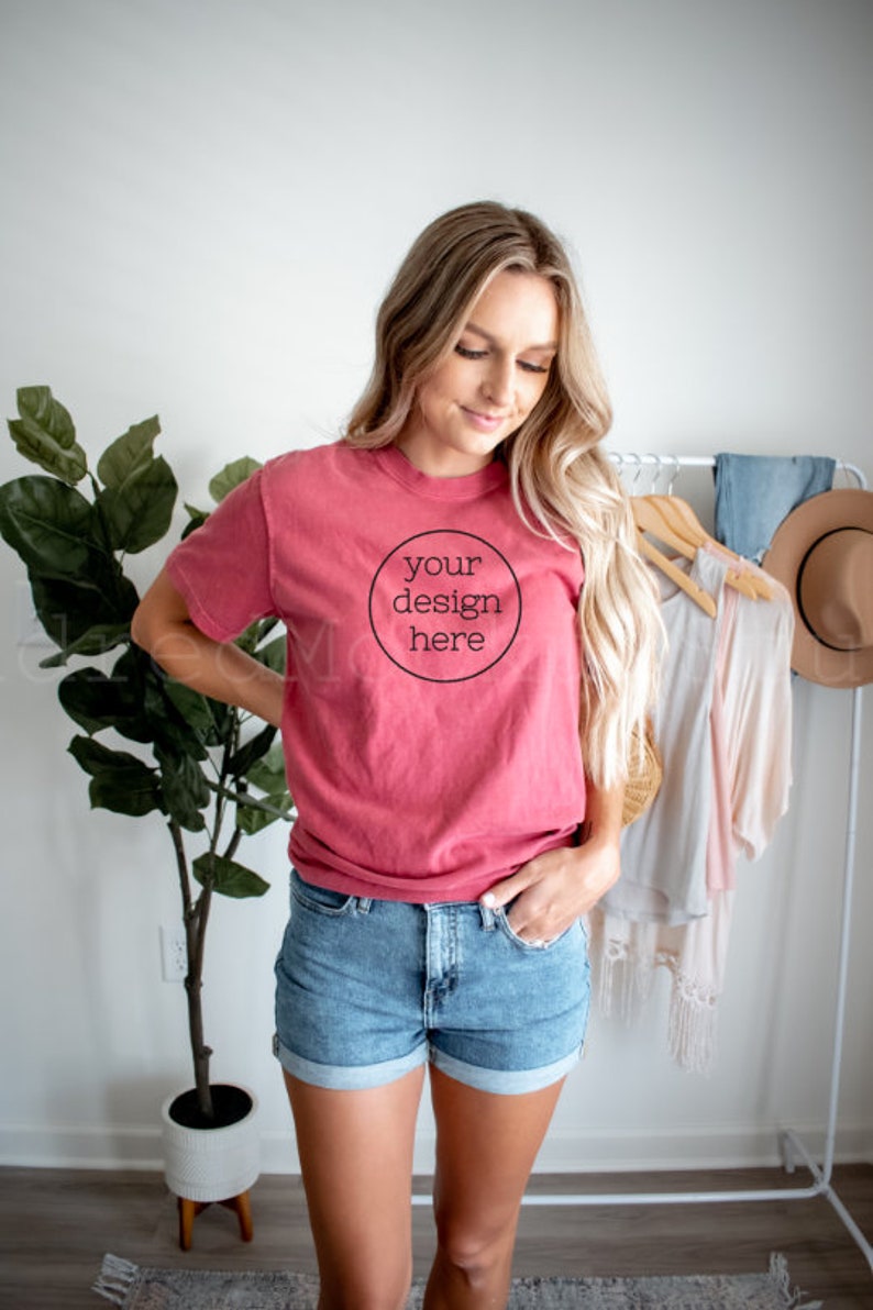 May include: A woman wearing a red t-shirt with the text "your design here" in a circle. She is wearing denim shorts and has her hand in her pocket.