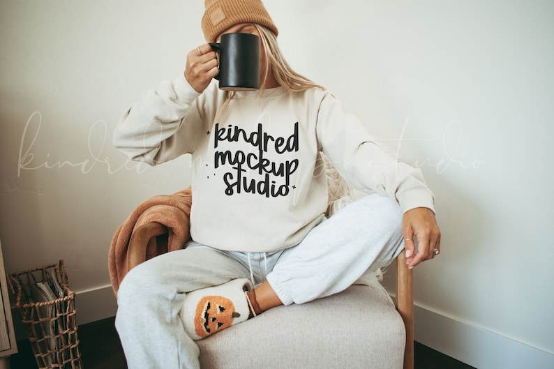 May include: A woman wearing a beige sweatshirt with the text "kindred mockup studio" in black lettering. She is sitting in a chair with a blanket on her lap and is holding a black mug.