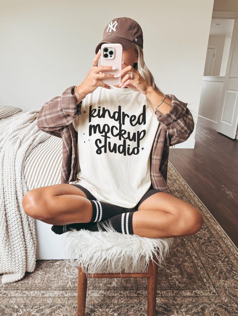 May include: A person wearing a brown baseball cap and a plaid shirt, holding a phone. The person is wearing a white t-shirt with the text "kindred mockup studio" in black script. The person is sitting on a wooden stool with a white fur seat, wearing black shorts and black socks with white stripes.