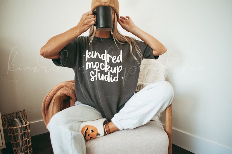 Pode incluir: Uma mulher vestindo uma camiseta cinza-chumbo com o texto "kindred mockup studio" em letras brancas. Ela est&aacute; sentada em uma cadeira com um cobertor marrom e segurando uma caneca preta.