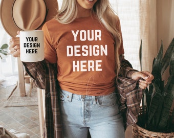Op de afbeelding: Een vrouw draagt een roestkleurig T-shirt met de tekst "YOUR DESIGN HERE" in wit. Ze houdt een witte mok vast met dezelfde tekst in zwart. Ze draagt ook een geruit overhemd en een blauwe jeans.