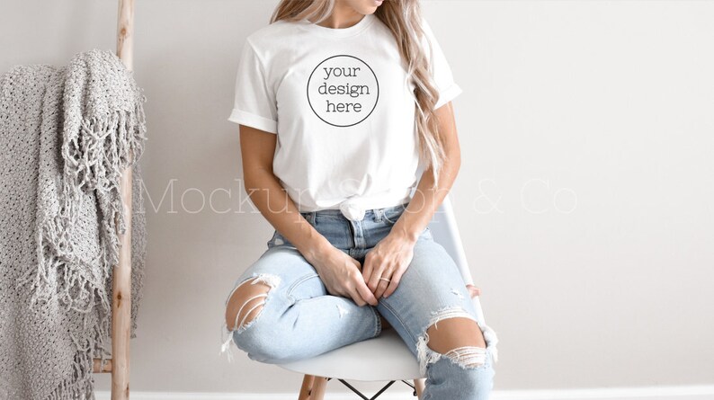 Pode incluir: Uma camiseta branca com um c&iacute;rculo na frente que diz "your design here". A camiseta est&aacute; sendo usada por uma pessoa sentada em uma cadeira branca. A pessoa est&aacute; usando jeans azul.