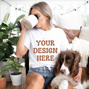 Puede incluir: Una mujer con una camiseta blanca con el texto "YOUR DESIGN HERE" en letras marrones, sentada en un patio con un perro. Lleva una taza blanca y gafas de sol.