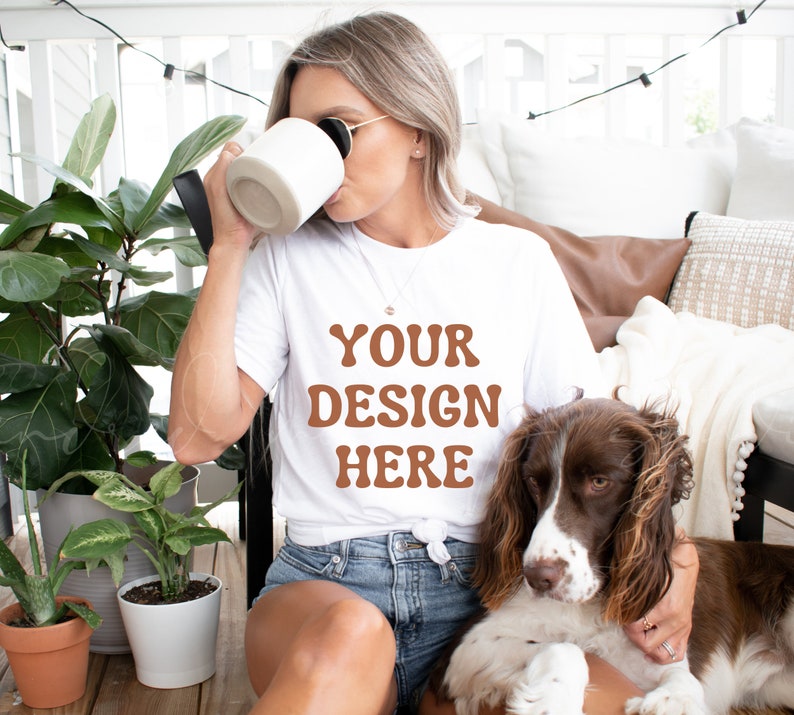 Puede incluir: Una mujer con una camiseta blanca con el texto "YOUR DESIGN HERE" en letras marrones, sentada en un patio con un perro. Lleva una taza blanca y gafas de sol.