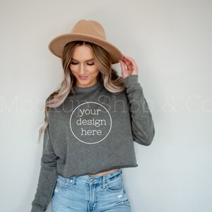 May include: A woman wearing a brown hat and a gray cropped sweatshirt with the text "your design here" in a circle. She is wearing blue jeans.