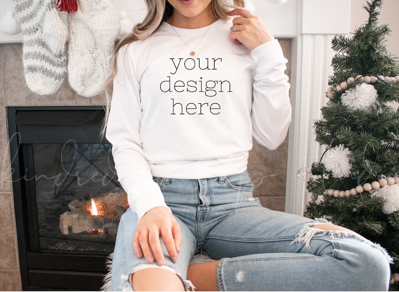 May include: White long-sleeve sweatshirt with the text "your design here" printed on the front. The sweatshirt is worn by a person sitting in front of a fireplace.
