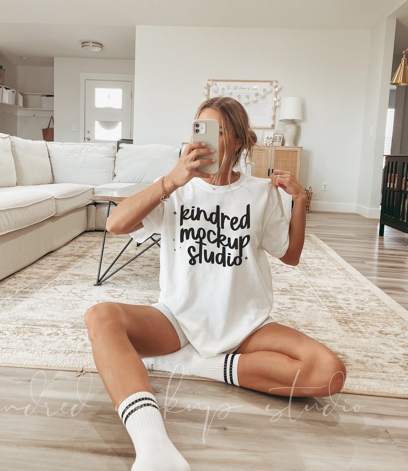Puede incluir: Una camiseta blanca con el texto "kindred mockup studio" en letras negras. La camiseta la lleva puesta una persona sentada en una alfombra.