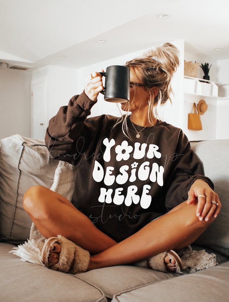 Pu&ograve; includere: Una donna indossa una felpa marrone con il testo "YOUR DESIGN HERE" in bianco. &Egrave; seduta su un divano e tiene una tazza nera.