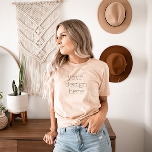 May include: A woman wearing a light peach t-shirt with the text "your design here" printed on the front. She is standing in front of a white wall with a macrame wall hanging and two brown hats.