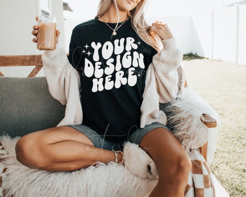 May include: A black t-shirt with the text "YOUR DESIGN HERE" in white, with a retro font style. The shirt is being worn by a person sitting on a couch.