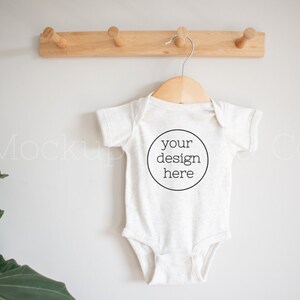May include: A white baby onesie with short sleeves hangs on a wooden hanger. The onesie has a black circle with the text "your design here" in the center.