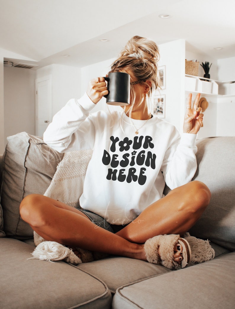 Pode incluir: Uma camisa de moletom branca com o texto "YOUR DESIGN HERE" em letras pretas de estilo retr&ocirc;. A camisa de moletom est&aacute; sendo usada por uma pessoa sentada em um sof&aacute;.