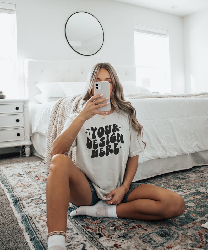 Puede incluir: Una camiseta blanca con el texto "YOUR DESIGN HERE" en letras negras de estilo retro. La camiseta la lleva puesta una persona sentada en una alfombra.