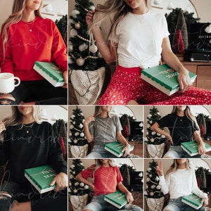 Op de afbeelding: Een collectie tops in verschillende kleuren, waaronder rood, wit, zwart, grijs en rood, gecombineerd met jeans. Elke persoon houdt een groene doos vast met de tekst "'Tis the Season". Een kerstboom en decoraties zijn op de achtergrond te zien.